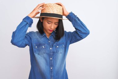 Genç ve güzel Çinli kadın kot gömlek ve şapka giyip izole edilmiş beyaz arka plan üzerinde acı ve migren yüzünden çaresiz ve stresli bir baş ağrısından muzdarip. Ellerini başının üstüne koy..