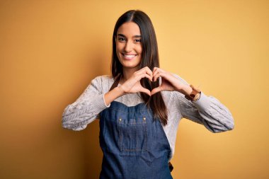 Sarı arka planda yemek pişiren önlük giymiş genç, güzel fırıncı kadın. Aşkla gülümsüyor. Elleriyle kalp sembolü ve şekli gösteriyor. Romantik konsept.