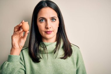 Young brunette woman with blue eyes holding fresh raw egg over isolated background with a confident expression on smart face thinking serious