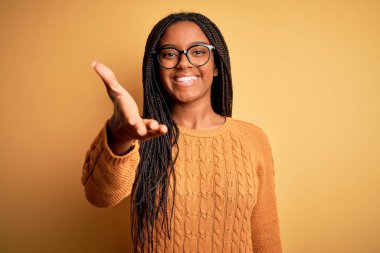Young african american smart woman wearing glasses and casual sweater over yellow background smiling friendly offering handshake as greeting and welcoming. Successful business.