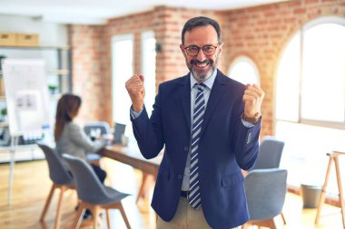 Middle age handsome businessman wearing glasses   standing at the office celebrating surprised and amazed for success with arms raised and open eyes. Winner concept.