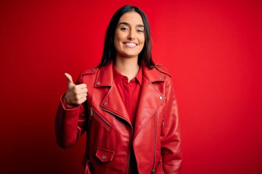Genç, güzel, esmer, sıradan bir ceket giyen, izole edilmiş kırmızı arka planda duran, mutlu baş parmaklı el hareketi yapan bir kadın. Kameraya bakıp başarıyı gösteren ifade onaylanıyor.