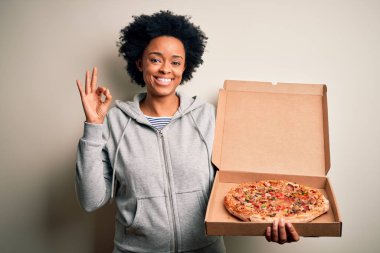 Young African American afro woman with curly hair holding delivery box with Italian pizza doing ok sign with fingers, excellent symbol