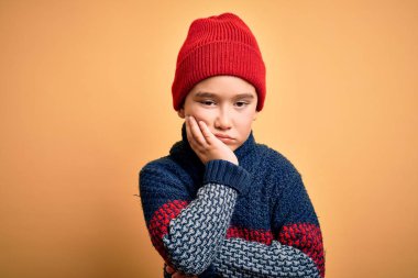 Young little boy kid wearing wool cap and winter sweater over yellow isolated background thinking looking tired and bored with depression problems with crossed arms.