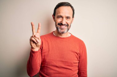Orta yaşlı, turuncu bir süveter giyen, izole edilmiş beyaz arka plan üzerinde duran, iki numarayı işaret eden, kendinden emin ve mutlu gülümseyen, orta yaşlı bir adam..