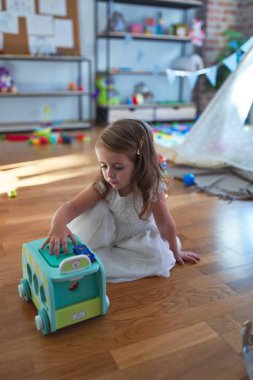 Anaokulunda bir sürü oyuncağın etrafında arabayla oynayan sevimli sarışın çocuk.