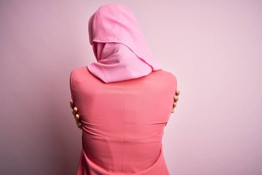 İslami tesettür takan genç, güzel bir kız pembe arka planda dikilip kendine mutlu ve pozitif bir şekilde sarılıyor. Sevgi ve özgüven.