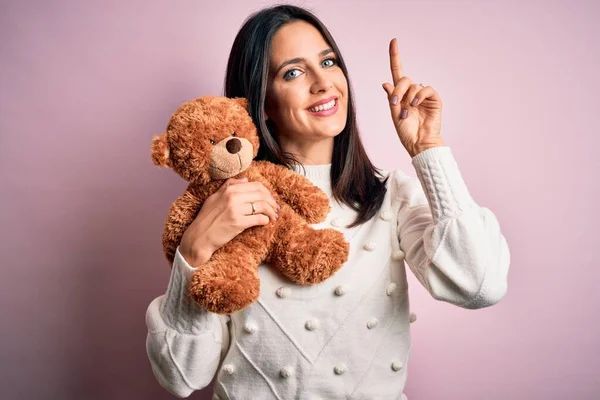 Young brunette woman with blue eyes hugging teddy bear stuffed animal over pink background surprised with an idea or question pointing finger with happy face, number one