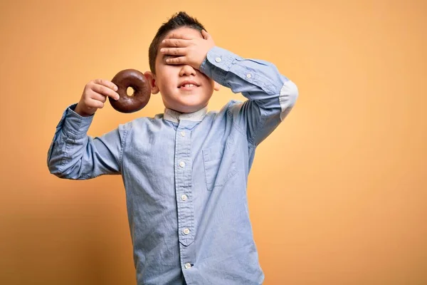 Młody Chłopiec Jedzący Niezdrowe Pączki Czekoladowe Odizolowanym Żółtym Tle Zestresowany — Zdjęcie stockowe
