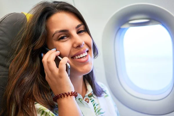 Havaalanında oturan genç yolcu kadın. Pencereden gökyüzü manzaralı. Akıllı telefon kullanarak arıyor.