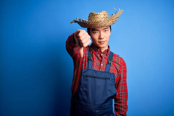 Young handsome chinese farmer man wearing apron and straw hat over blue background looking unhappy and angry showing rejection and negative with thumbs down gesture. Bad expression.