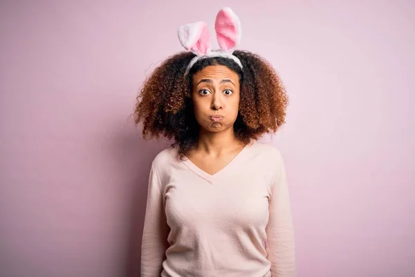 ピンクの背景にウサギの耳を身に着けているアフロの髪を持つ若いアフリカ系アメリカ人女性は面白い顔で頬をパフ 口は空気で膨脹し 狂気の表現 — ストック写真