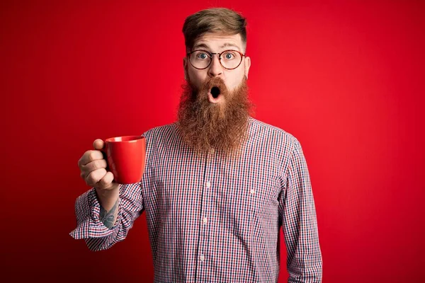 红头发爱尔兰男人 留着胡子 在红色背景下喝着一杯热咖啡 吓得满脸诧异 吓得面无血色 吓得面无血色 — 图库照片