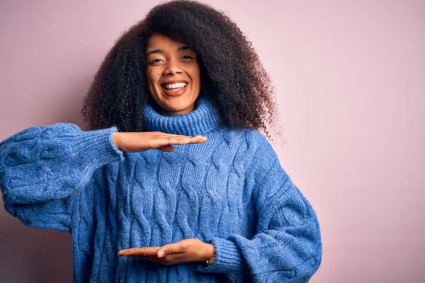 Young beautiful african american woman with afro hair wearing winter sweater over pink background gesturing with hands showing big and large size sign, measure symbol. Smiling looking at the camera. Measuring concept.