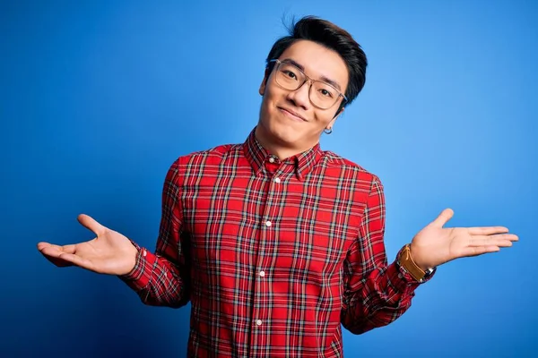 Young handsome chinese man wearing casual shirt and glasses over blue background clueless and confused expression with arms and hands raised. Doubt concept.