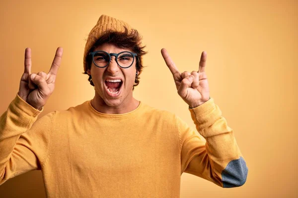 孤立した黄色の背景にカジュアルなTシャツと眼鏡を身に着けている若いハンサムな男が手を上げて岩のシンボルをやって狂気の表情で叫んでいます 音楽スター 重い概念 — ストック写真