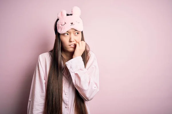 年轻的亚洲女人戴着睡衣 戴着睡眠面罩 披着粉色的隔离背景 看上去很紧张 双手放在咬指甲的嘴上 焦虑问题 — 图库照片