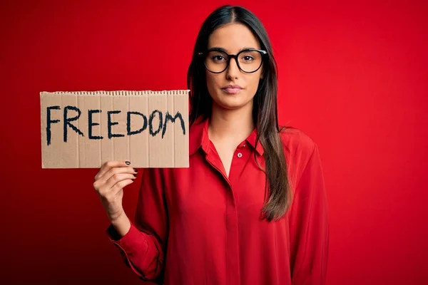 Young beautiful brunette activist woman wearing glasses protesting for freedom holding poster with a confident expression on smart face thinking serious