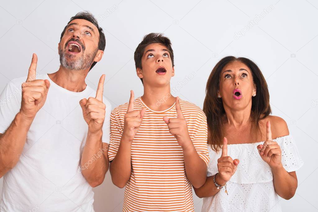 Family of three, mother, father and son standing over white isolated background amazed and surprised looking up and pointing with fingers and raised arms.