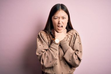 Güzel Asyalı bir kadın pembe arka planda şık bir süveter giyip boğularak can çekişiyor çünkü boğularak ölüyor. Sağlık problemi. Boğulma ve intihar konsepti.