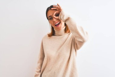 Beautiful redhead woman wearing winter turtleneck sweater over isolated background doing ok gesture with hand smiling, eye looking through fingers with happy face.
