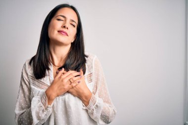 Young brunette woman with blue eyes wearing casual t-shirt over isolated white background smiling with hands on chest with closed eyes and grateful gesture on face. Health concept.