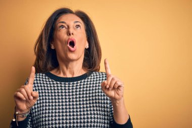 Middle age beautiful woman wearing casual sweater over isolated yellow background amazed and surprised looking up and pointing with fingers and raised arms.