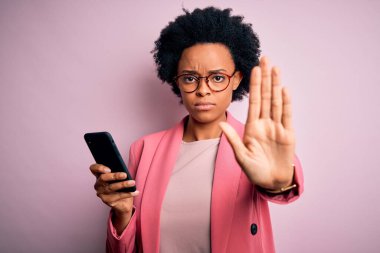 Young African American afro woman with curly hair having conversation using smartphone with open hand doing stop sign with serious and confident expression, defense gesture