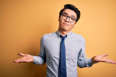 Genç yakışıklı Çinli iş adamı gözlük takıyor ve sarı arka plan üzerinde kravat takıyor. Kolları ve elleri havada, kafası karışmış bir ifade takınıyor. Şüphe kavramı.