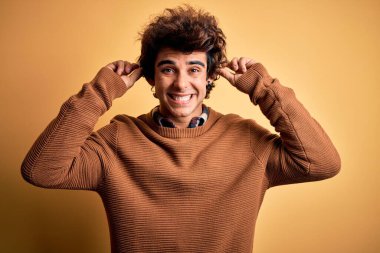 İzole edilmiş sarı arka planda sıradan tişört ve süveter giyen genç yakışıklı adam parmaklarıyla kulaklarını çekerek gülüyor, komik bir jest yapıyor. Seçme sorunu