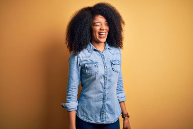 Afro saçlı, güzel, Afro-Amerikan bir kadın sarı izole edilmiş arka planda duruyor. Seksi bir ifadeyle kameraya bakıyor, neşeli ve mutlu bir yüzle..