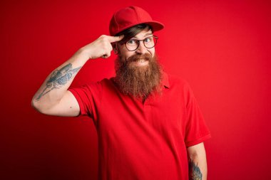 Gözlüklü ve kırmızı şapkalı genç teslimatçı izole edilmiş arka planda gülümsüyor. Tek parmakla, harika bir fikir ya da düşünceyle, iyi bir hafızayla.