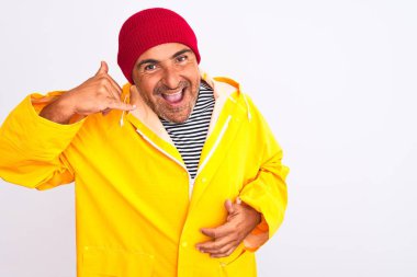 Middle age man wearing rain coat and woolen hat standing over isolated white background smiling doing phone gesture with hand and fingers like talking on the telephone. Communicating concepts.