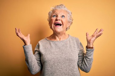Senior beautiful woman wearing casual t-shirt standing over isolated yellow background celebrating mad and crazy for success with arms raised and closed eyes screaming excited. Winner concept