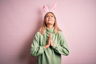 Paskalya tavşanı kulakları takan genç, güzel bir kadın pembe arka planda duruyor. Elleriyle yalvarıyor ve dua ediyor. Yüzünde umut ifadesi var. Yalvarıyorum..