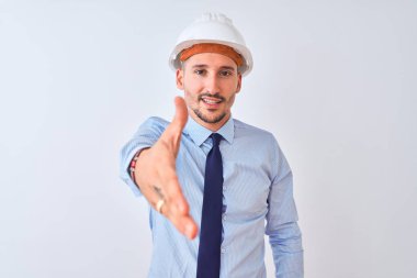 Young business man wearing contractor safety helmet over isolated background smiling friendly offering handshake as greeting and welcoming. Successful business.