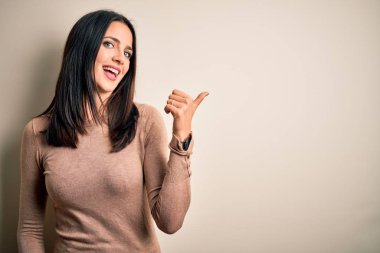 Young brunette woman with blue eyes wearing casual sweater over isolated white background smiling with happy face looking and pointing to the side with thumb up.