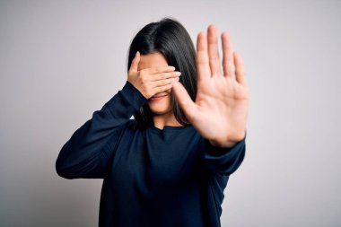 Young beautiful brunette woman wearing casual sweater standing over white background covering eyes with hands and doing stop gesture with sad and fear expression. Embarrassed and negative concept.