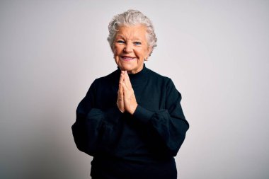 Senior beautiful woman wearing casual black sweater standing over isolated white background praying with hands together asking for forgiveness smiling confident.