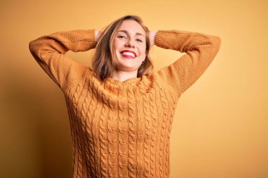 Sarı arka planda dikilen, kollarını ve ellerini başının ve boynunun arkasına koyup mutlu bir şekilde gülümseyen, güzel sarışın kadın.