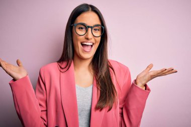 Young beautiful brunette businesswoman wearing jacket and glasses over pink background celebrating crazy and amazed for success with arms raised and open eyes screaming excited. Winner concept