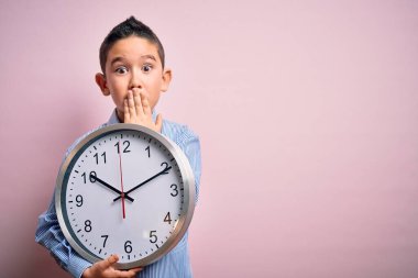 Young little boy kid holding big minute clock over isolated pink background cover mouth with hand shocked with shame for mistake, expression of fear, scared in silence, secret concept
