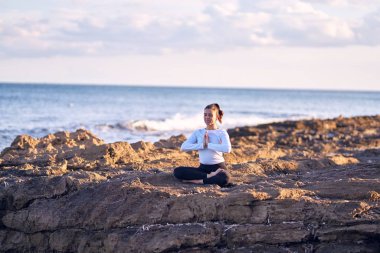 Genç ve güzel sporcu gülümseyerek yoga yapıyor. Yüzünde gülümseme olan koç sahilde dua öğretirken poz veriyor.