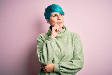 Young beautiful woman with blue fashion hair wearing casual turtleneck sweater with hand on chin thinking about question, pensive expression. Smiling with thoughtful face. Doubt concept.