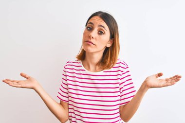 Beautiful redhead woman wearing casual striped pink t-shirt over isolated background clueless and confused expression with arms and hands raised. Doubt concept.