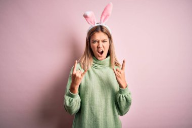 Paskalya tavşanı kulakları takan genç ve güzel bir kadın izole edilmiş pembe arka planda durup çılgın bir ifadeyle elleri havada kaya sembolü yaparak bağırıyor. Müzik yıldızı. Ağır konsept.