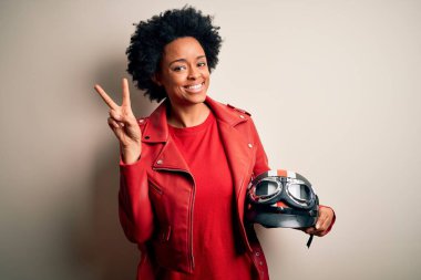 Young African American afro motorcyclist woman with curly hair holding motorcycle helmet smiling with happy face winking at the camera doing victory sign with fingers. Number two.
