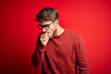 Young handsome man with beard wearing glasses and sweater standing over red background feeling unwell and coughing as symptom for cold or bronchitis. Health care concept.