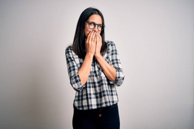 Young brunette woman with blue eyes wearing casual shirt and glasses over white background laughing and embarrassed giggle covering mouth with hands, gossip and scandal concept