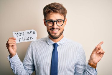 Young businessman wearing glasses holding paper with you are fired message very happy pointing with hand and finger to the side